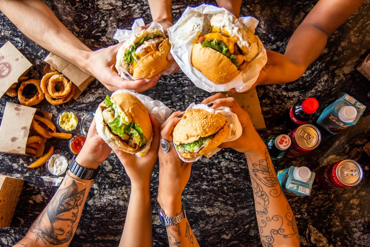Overhead view of four people holding burgers over a table of fries, onion rings and drinks.